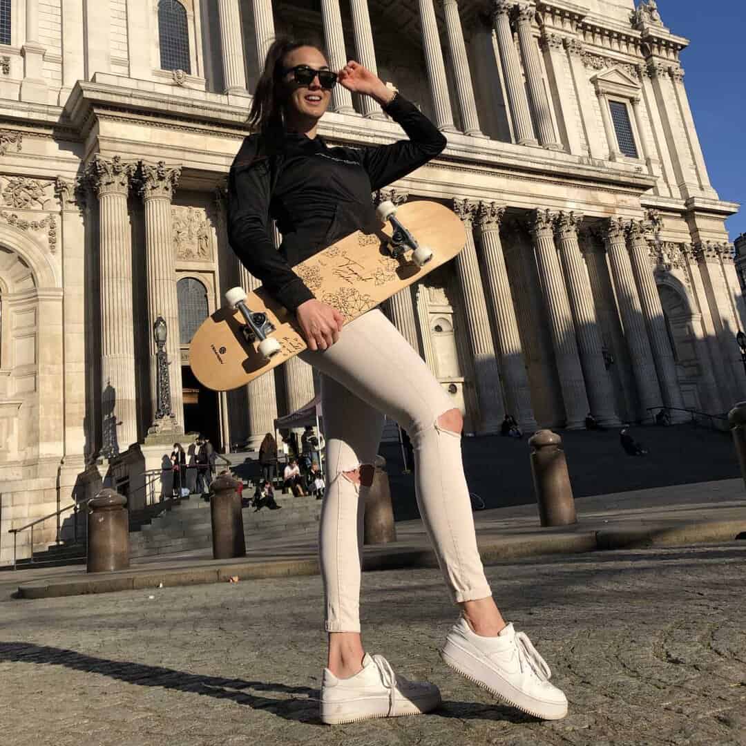 Жена с черна спортна екипировка и бели дънки, държаща скейтборд пред колоните на историческо здание в слънчев ден.